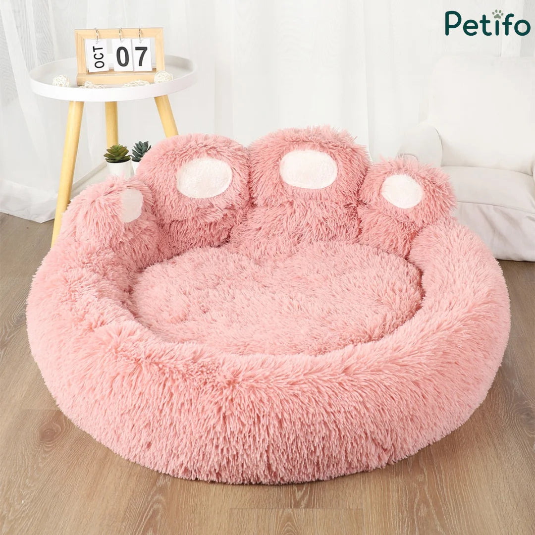 An empty pink fluffy paw-shaped dog bed displayed in a bright room with a small side table.