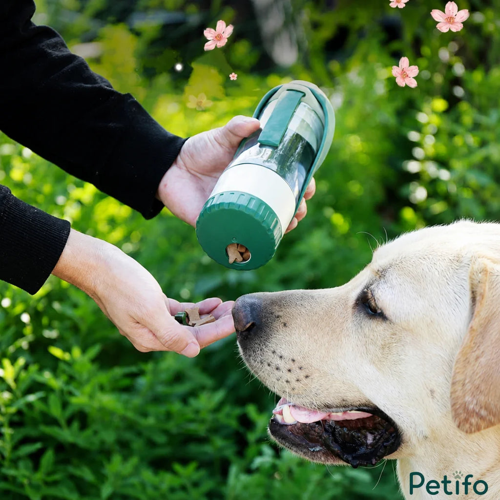 Person dispensing treats from the bottom compartment of a green Petifo portable water bottle next to a happy dog outdoors.