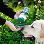 Person dispensing treats from the bottom compartment of a green Petifo portable water bottle next to a happy dog outdoors.