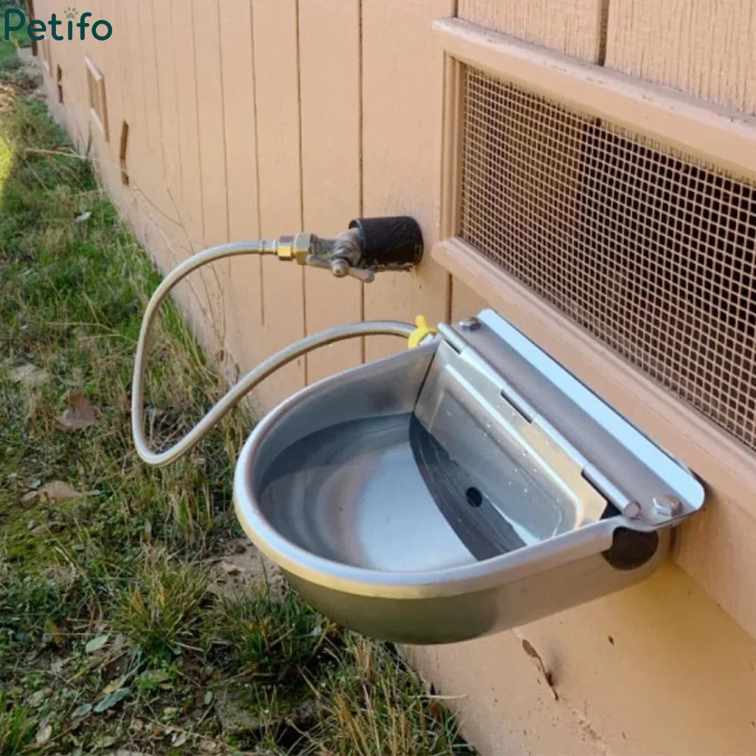 Fully installed automatic stainless steel livestock water bowl connected to an outdoor water supply line against a wall.