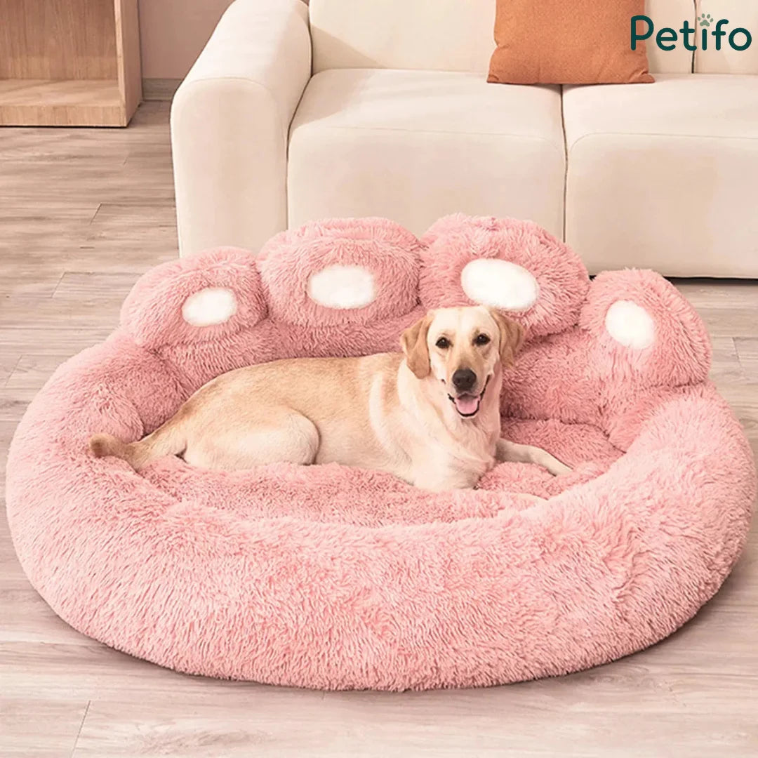 A golden retriever lying relaxed in a soft pink paw-shaped plush dog bed near a white couch.