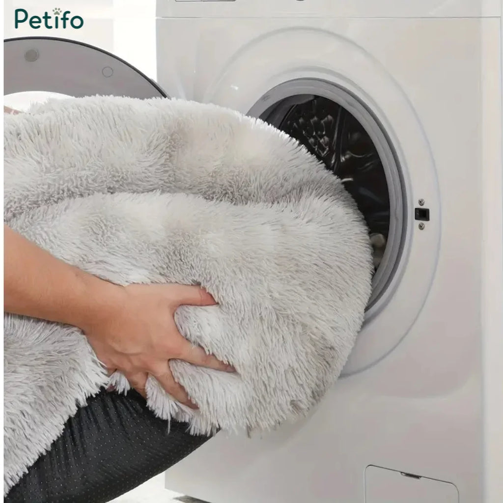 Person putting a fluffy grey pet calming bed into a washing machine.