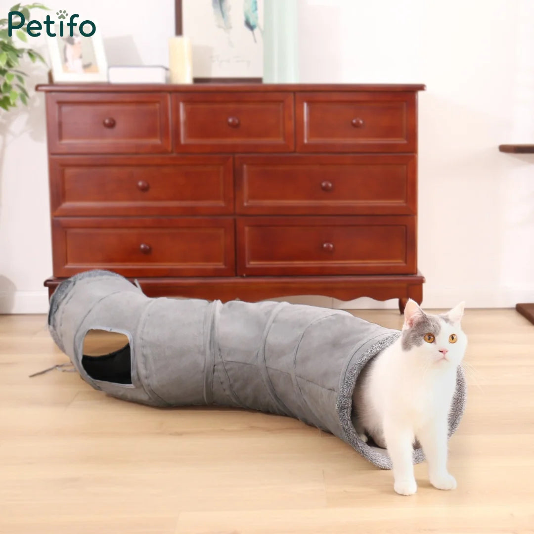 White and grey cat emerging from a collapsible grey plush cat tunnel on wooden flooring.